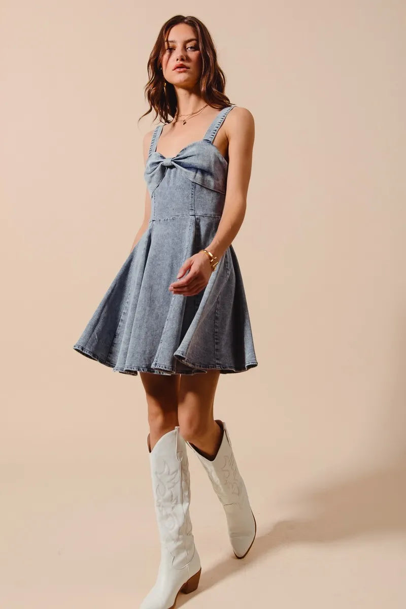 Woman wearing a denim dress with a bow tie neckline and white cowboy boots on a beige background