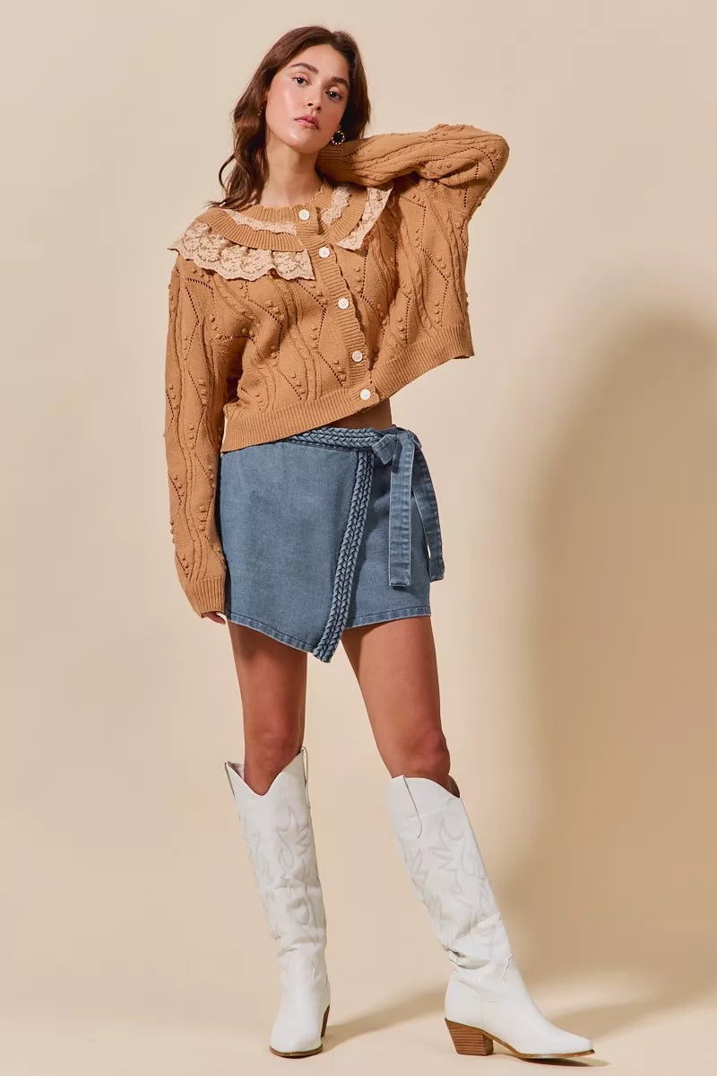 Woman wearing a brown cable knit sweater, denim skirt, and white knee-high boots on a beige background