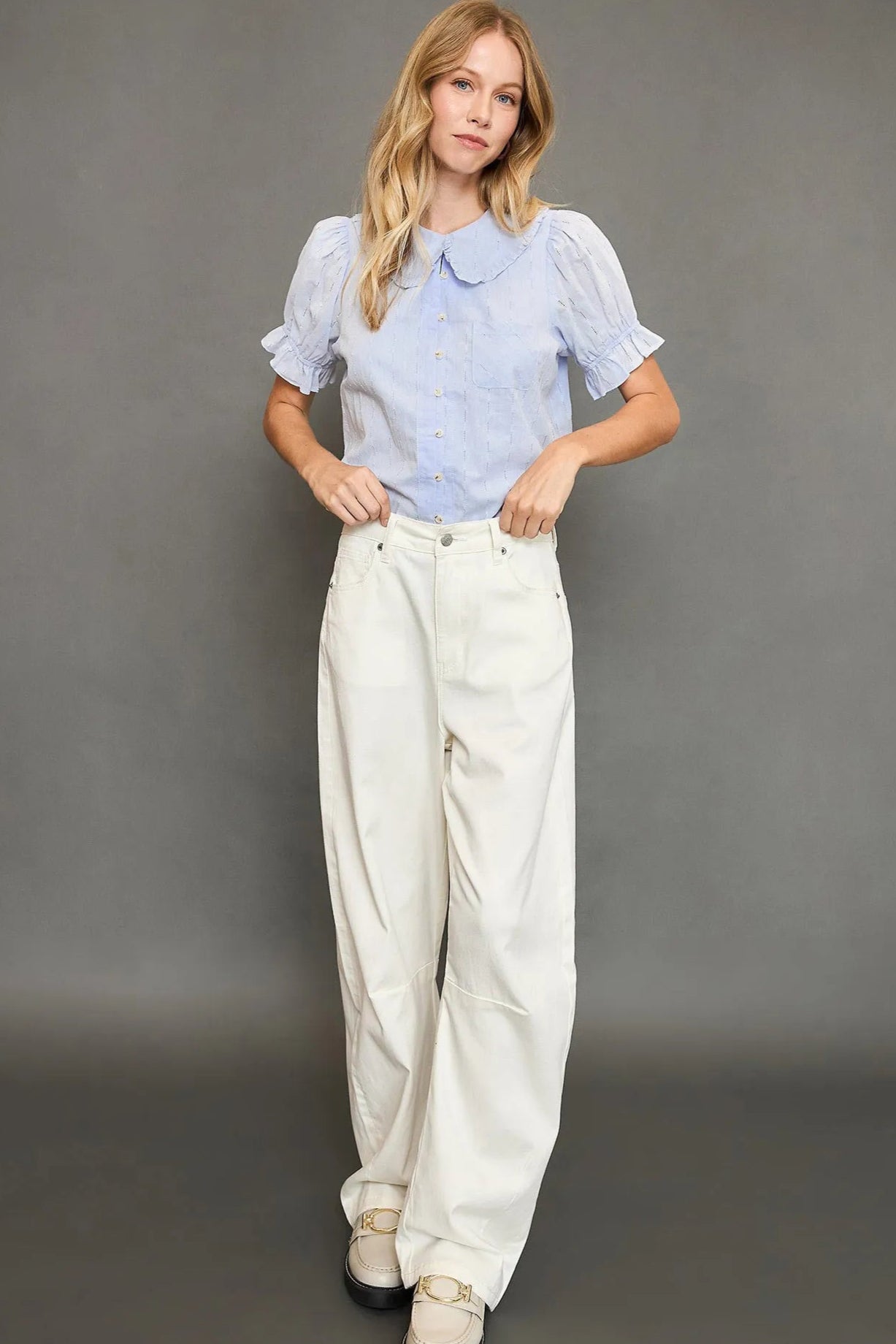 Woman wearing a light blue blouse and white pants against a gray background