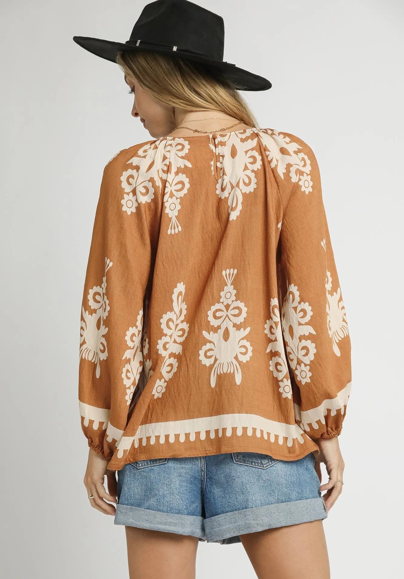 Person wearing a brown embroidered blouse with white patterns, denim shorts, and a black hat on a gray background.