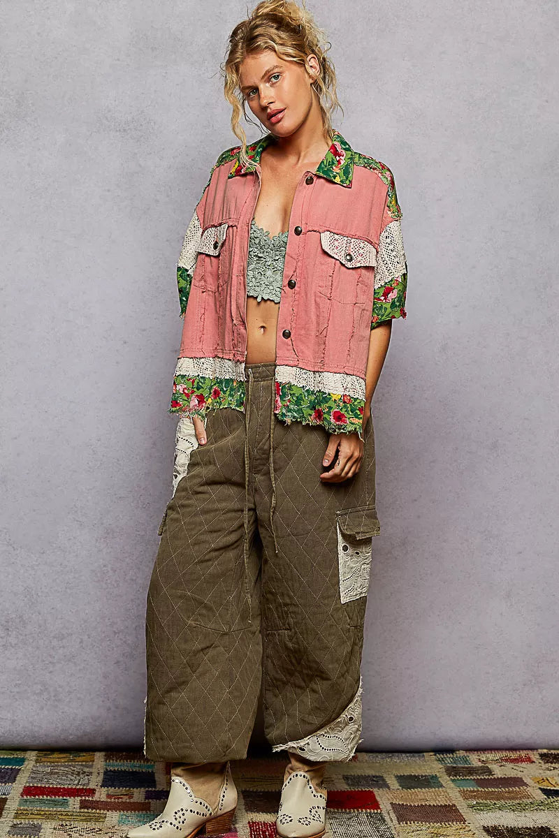 Woman wearing a colorful jacket and green pants against a gray wall.