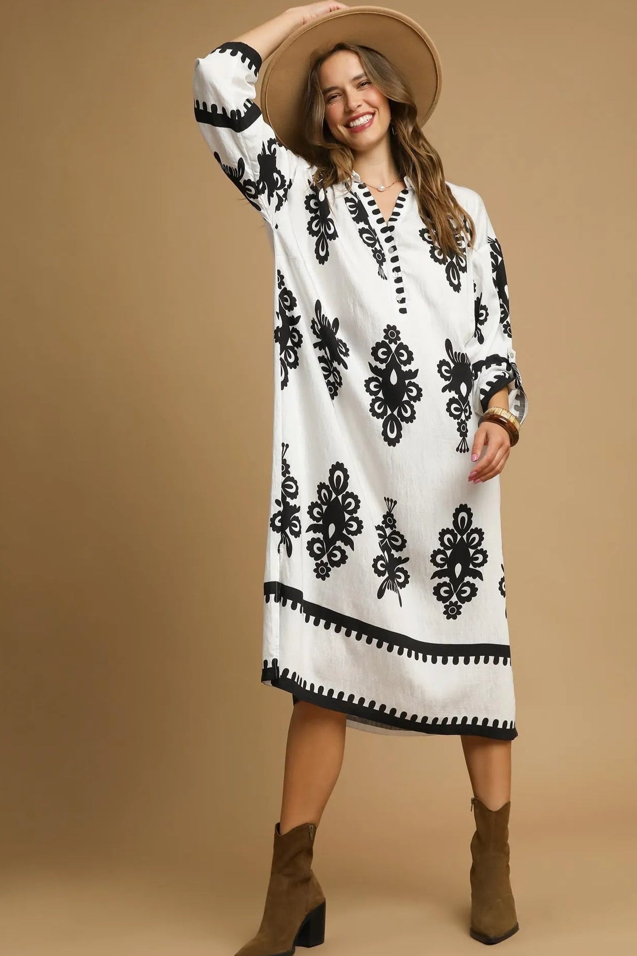 Woman wearing a white dress with black patterns, brown boots, and a wide-brimmed hat on a beige background.