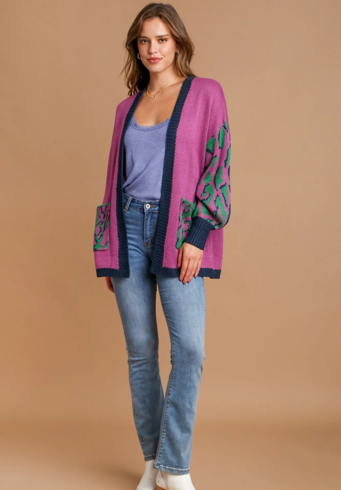Woman wearing a pink cardigan with green patterns, blue jeans, and white shoes on a beige background.