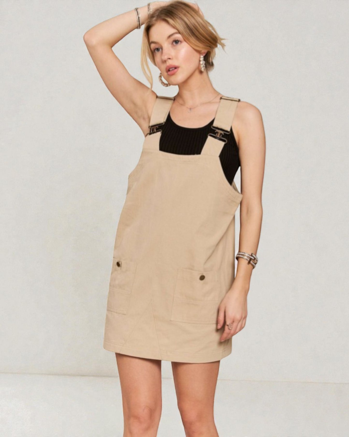 Woman wearing a beige pinafore dress with black top against a plain background