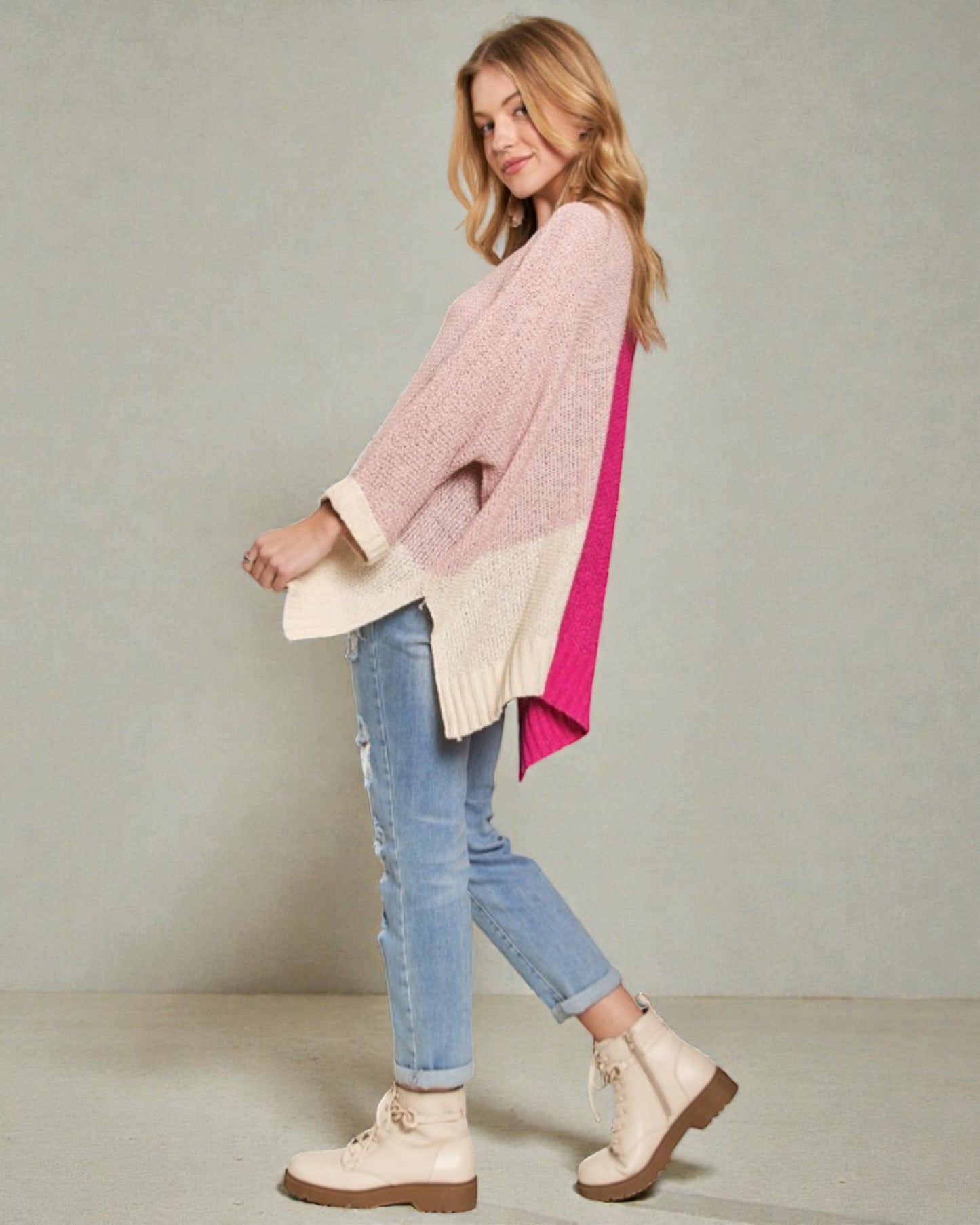 Woman wearing a pink and beige sweater with blue jeans and white boots on a plain background