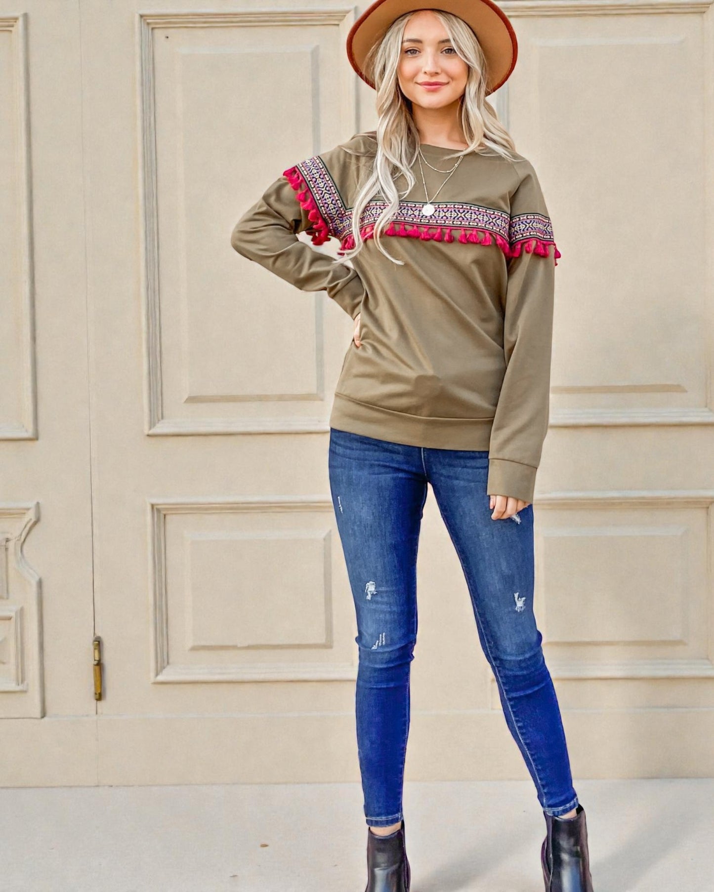 Woman wearing a green sweater with red accents and blue jeans standing in front of a beige wall.