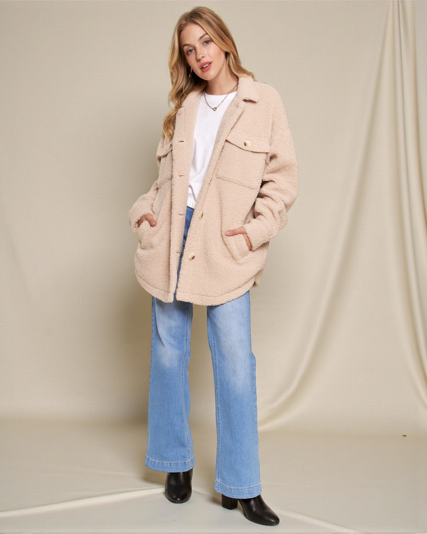 Woman wearing a beige coat, white shirt, blue jeans, and black shoes on a plain background