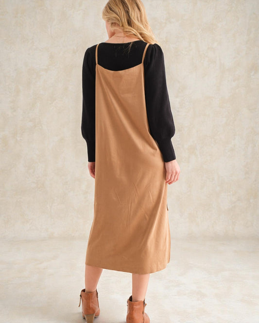 Woman wearing a black long-sleeve top and beige pinafore dress against a neutral background