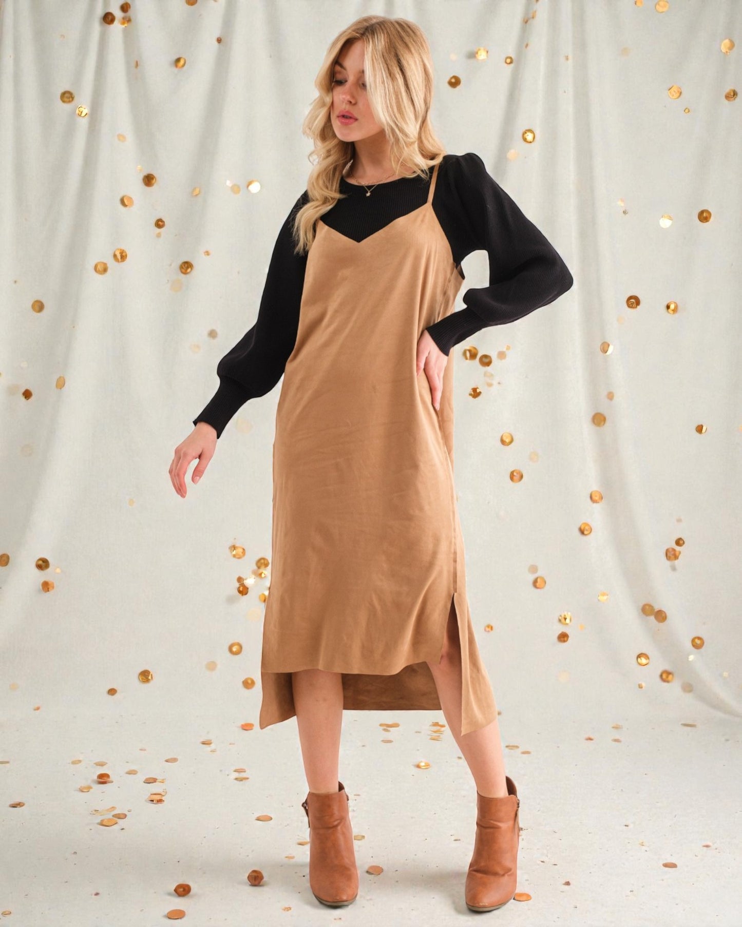 Woman wearing a beige pinafore dress with a black long-sleeve top against a light background with gold dots.