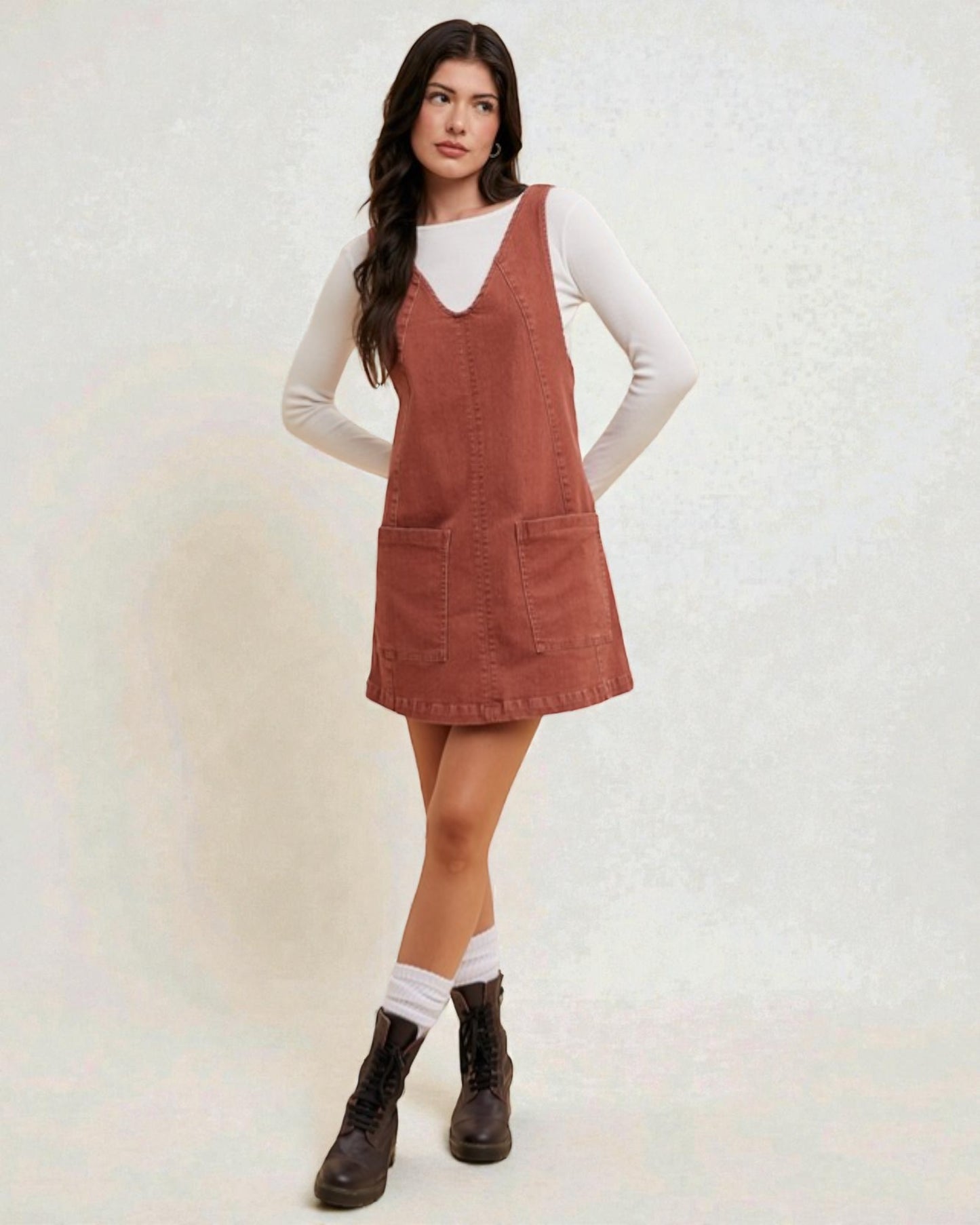 Woman wearing a brown pinafore dress over a white shirt on a light background