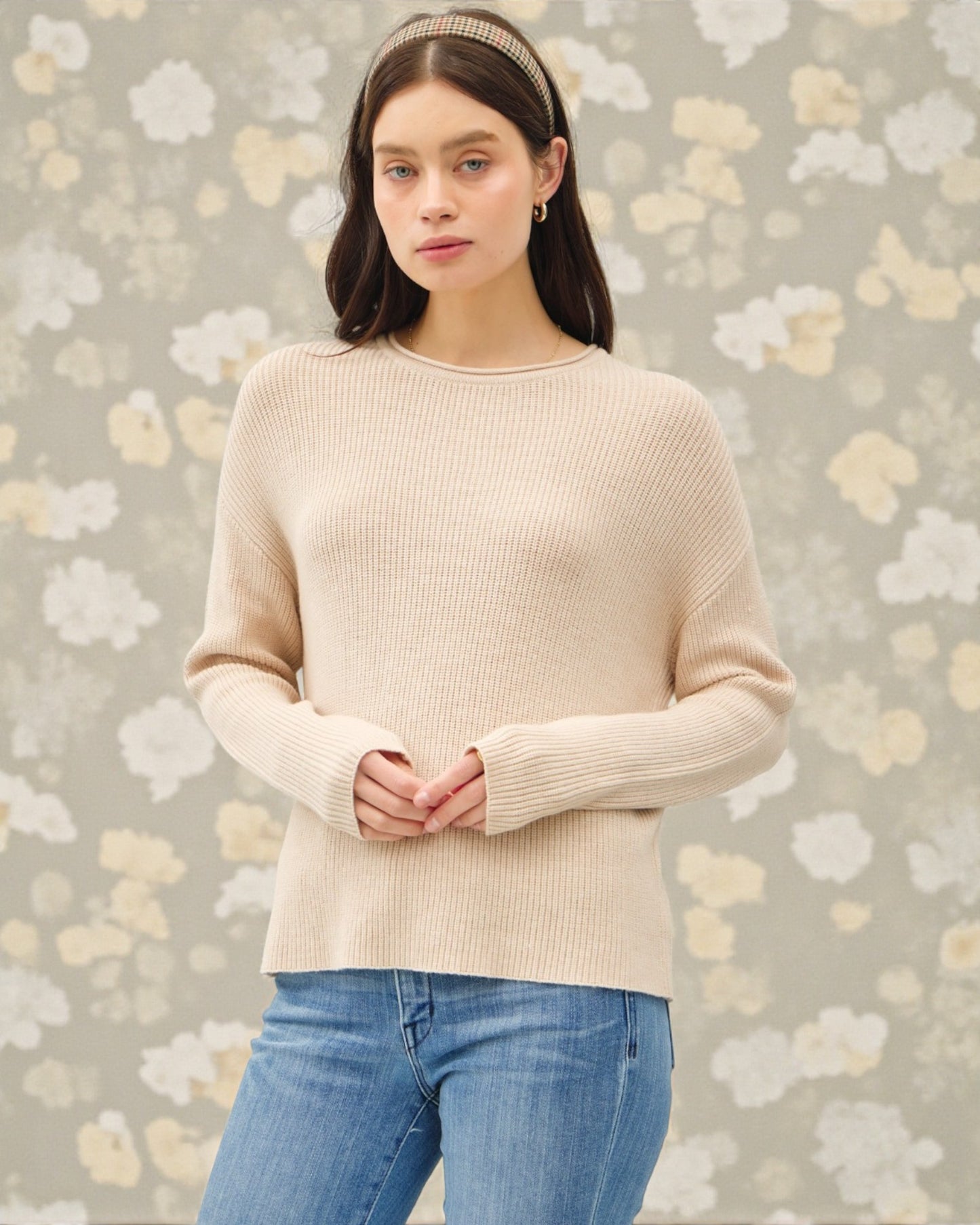 Woman wearing a beige sweater and blue jeans against a floral-patterned background