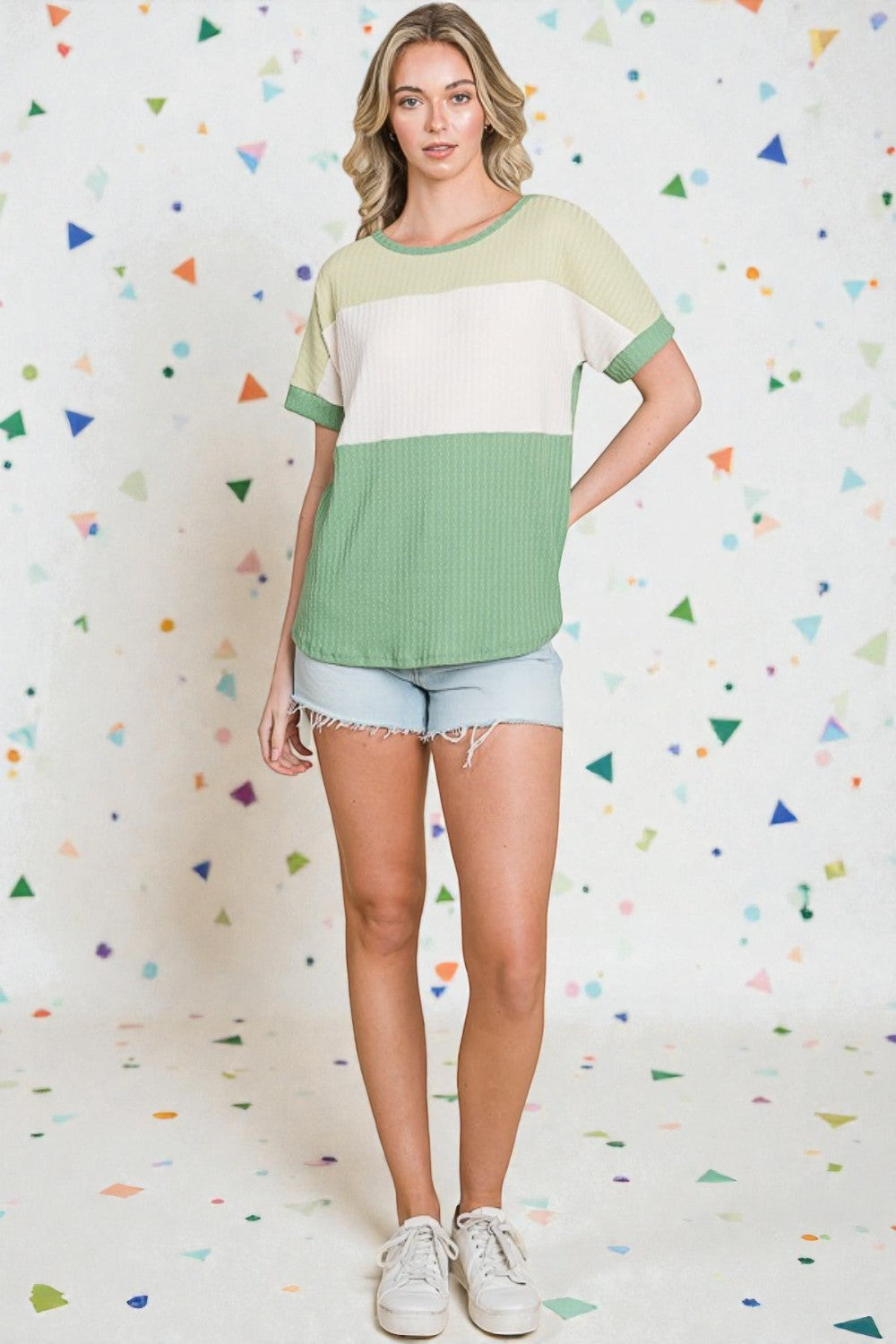 Woman wearing a green and white top with denim shorts against a colorful geometric-patterned wall.