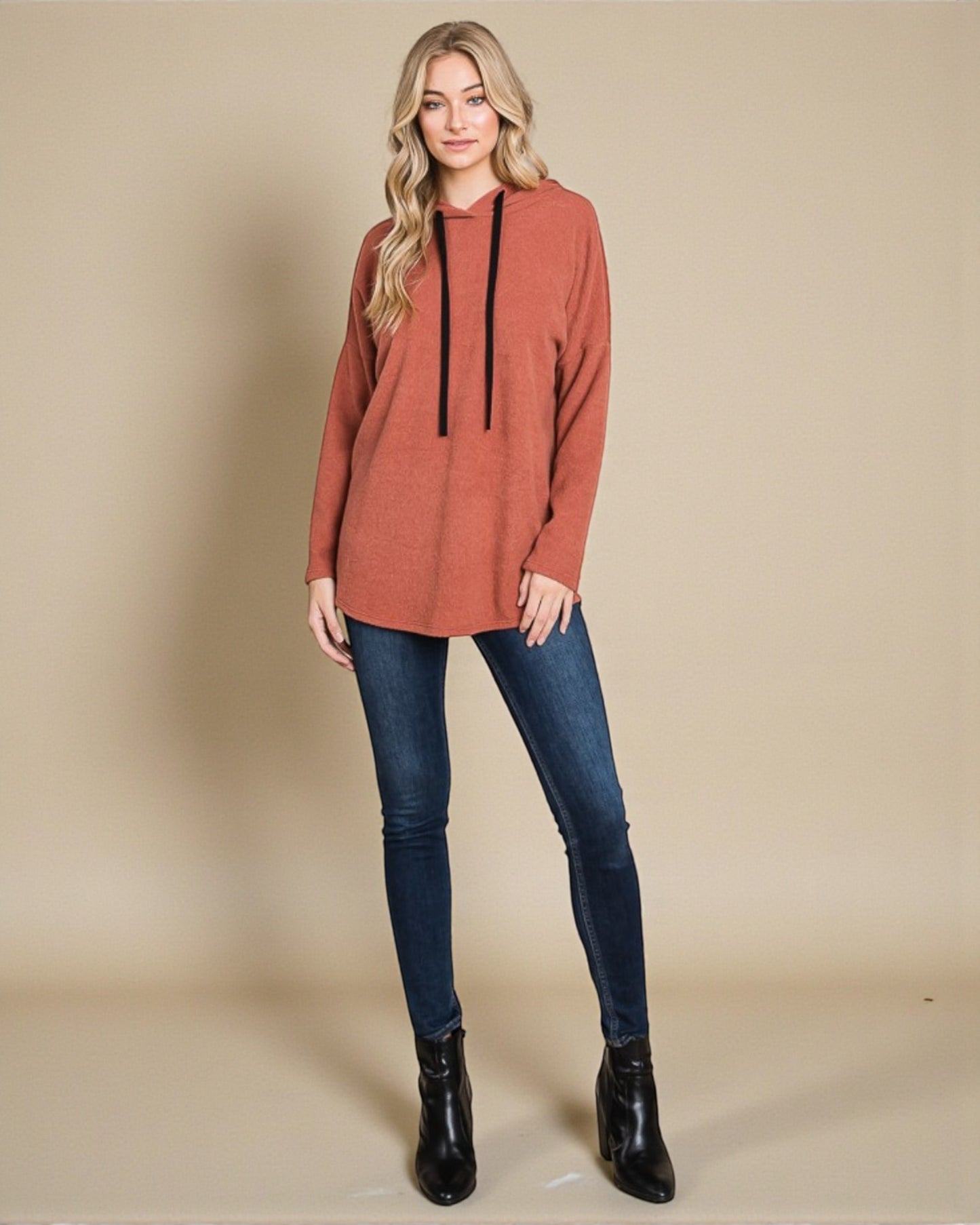 Woman wearing a rust-colored long-sleeve top with black accents, blue jeans, and black boots on a beige background.
