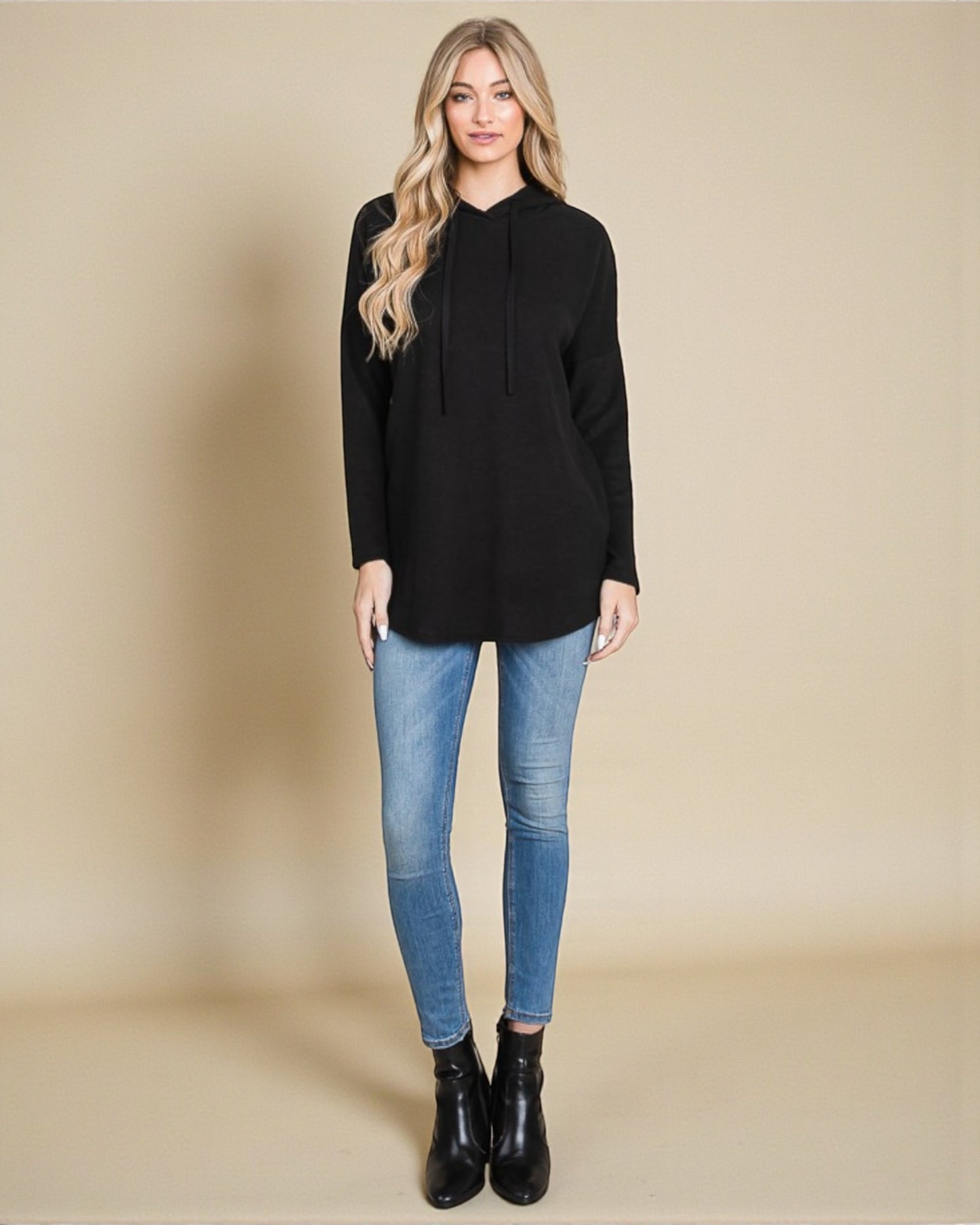 Woman wearing a black hoodie, blue jeans, and black boots on a beige background