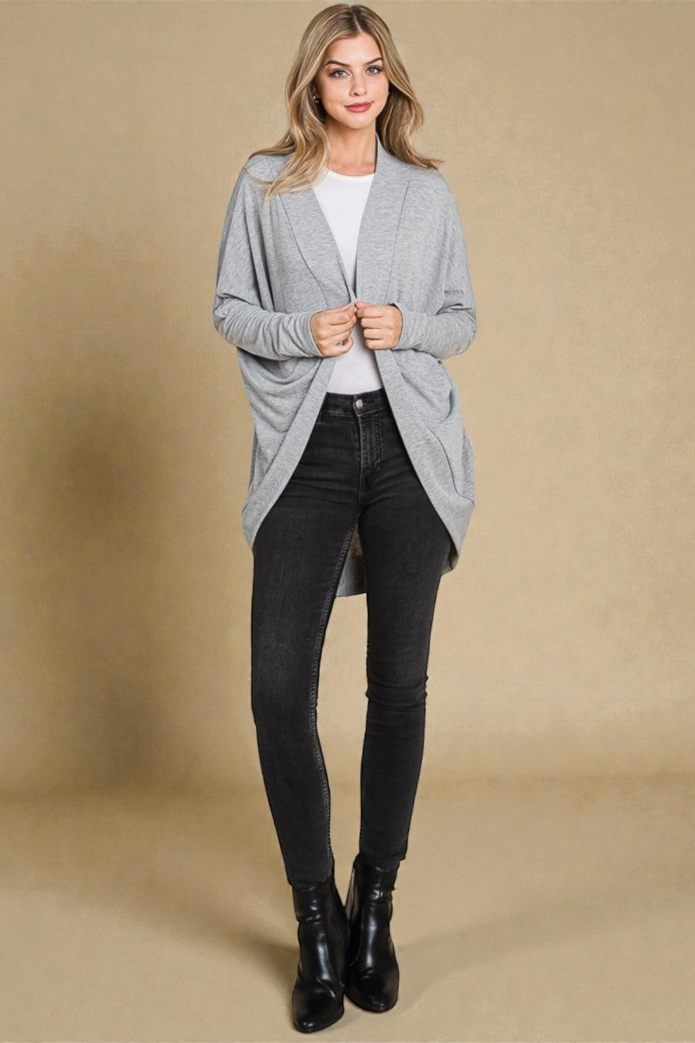 Woman wearing a gray cardigan, white shirt, black jeans, and black boots on a beige background