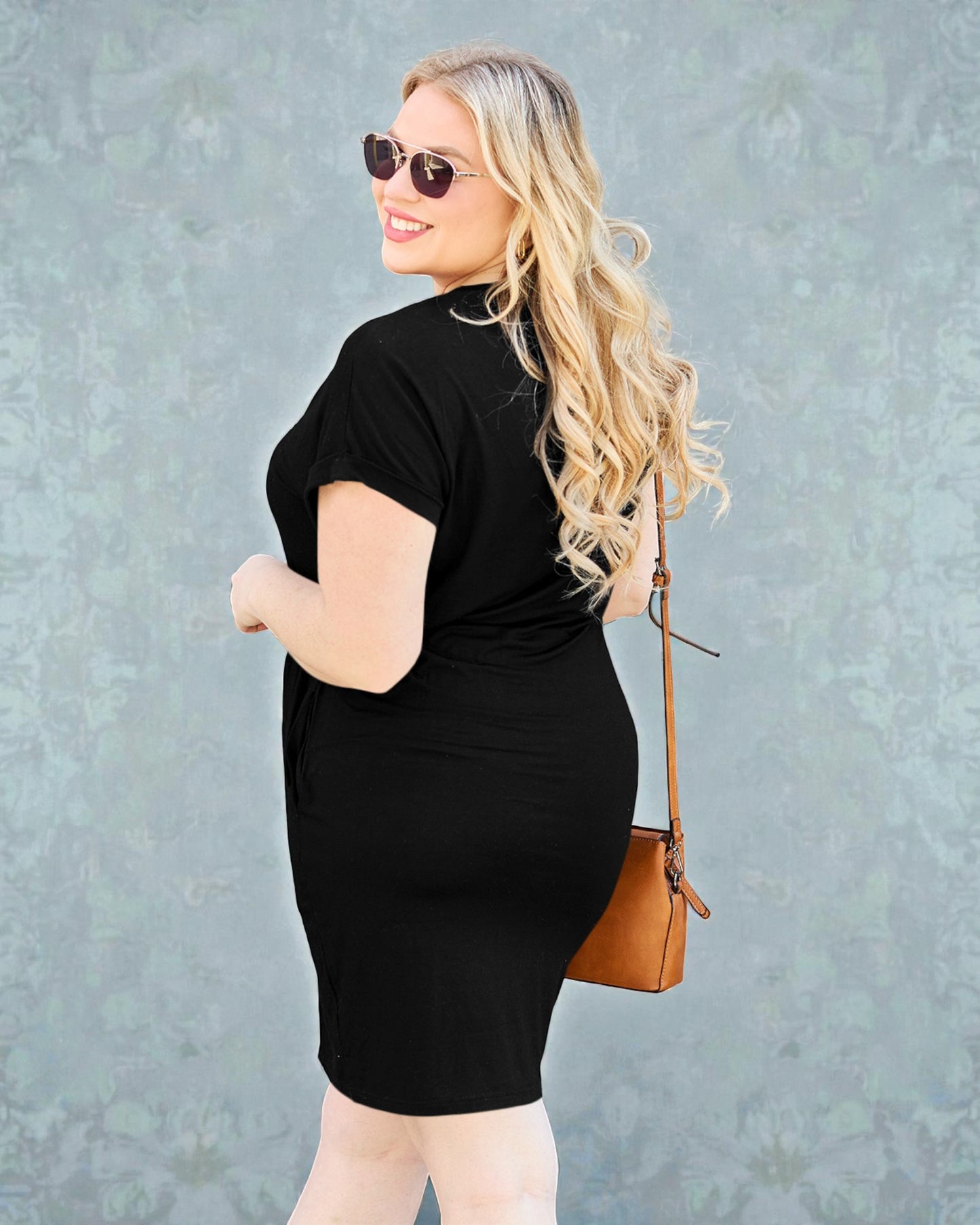 Woman wearing a black dress with sunglasses and a brown handbag against a gray background