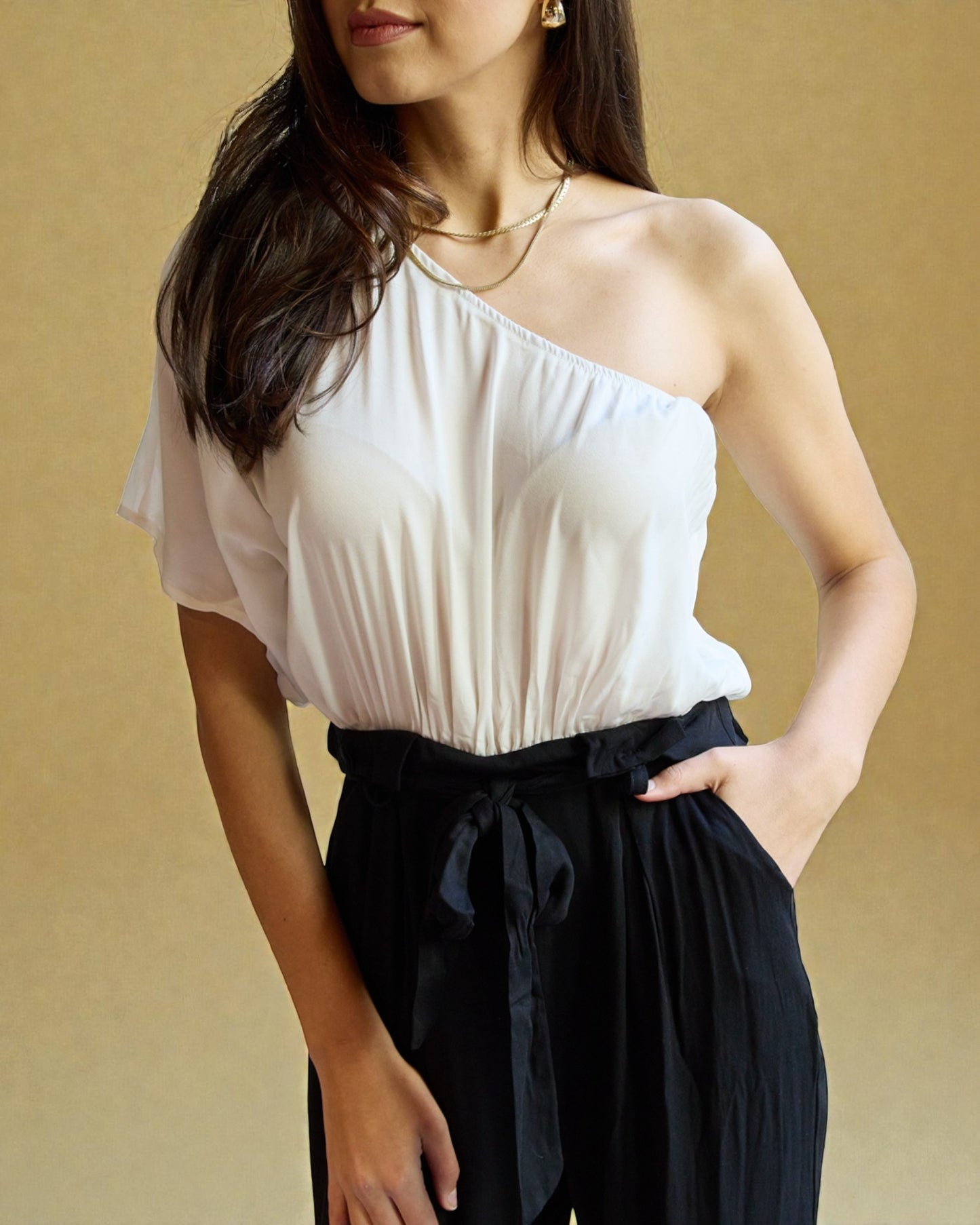 Woman wearing a white one-shoulder top and black pants against a beige background