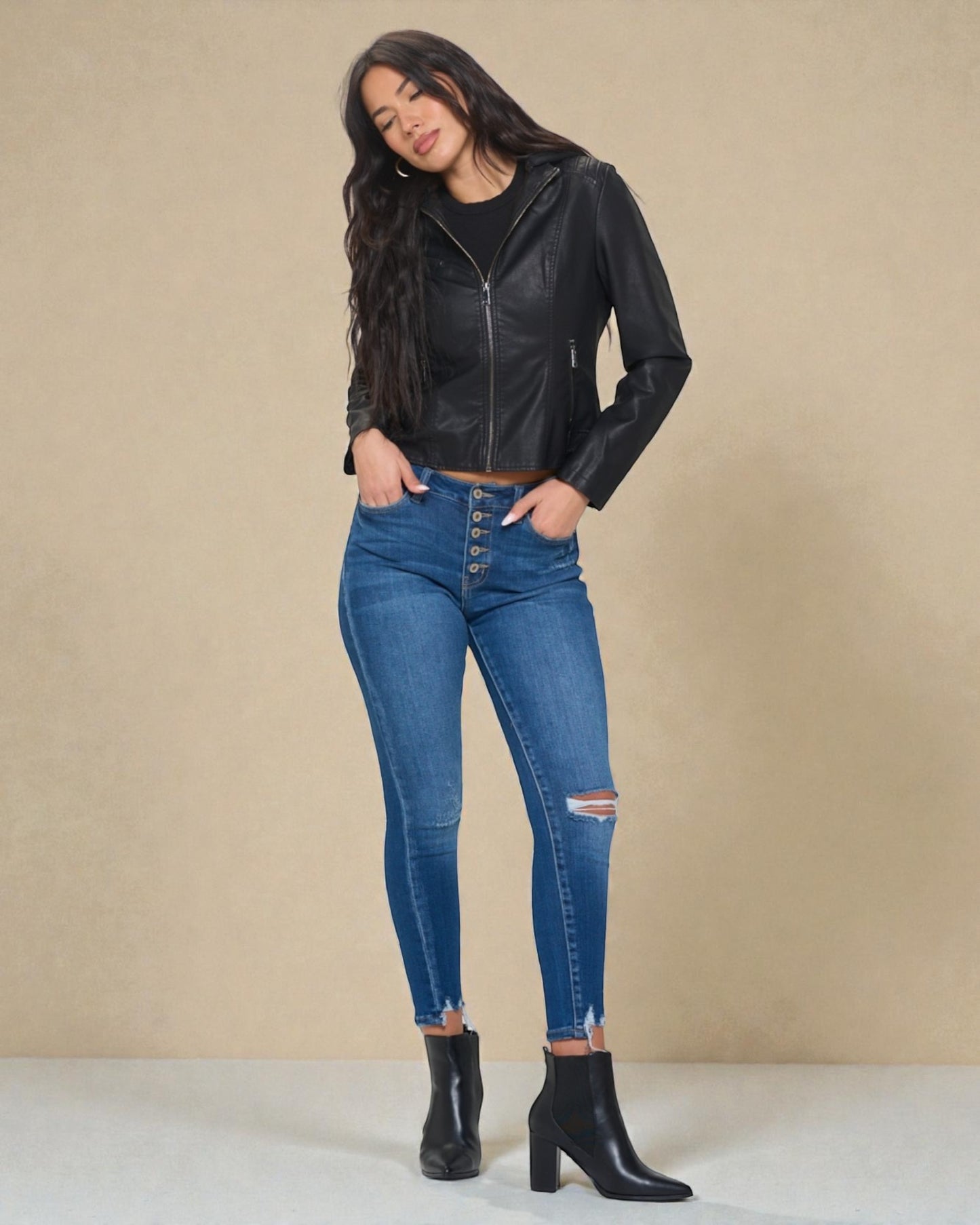 Woman wearing a black leather jacket, blue jeans, and black boots on a beige background