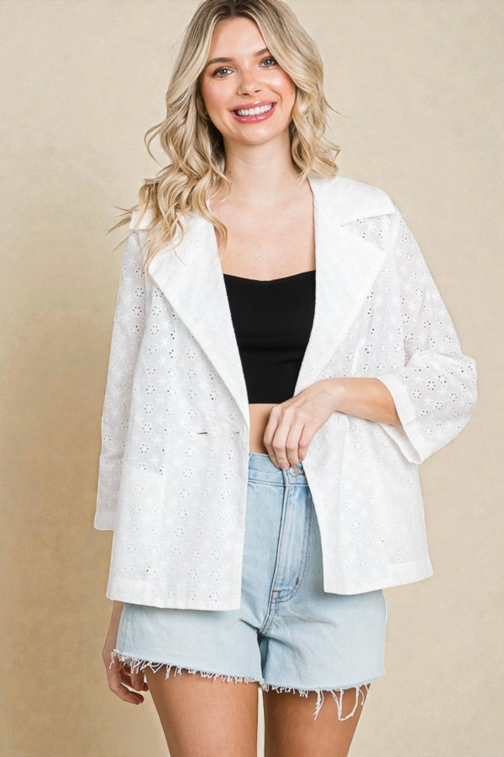 Woman wearing a white lace jacket over a black top and denim shorts on a beige background