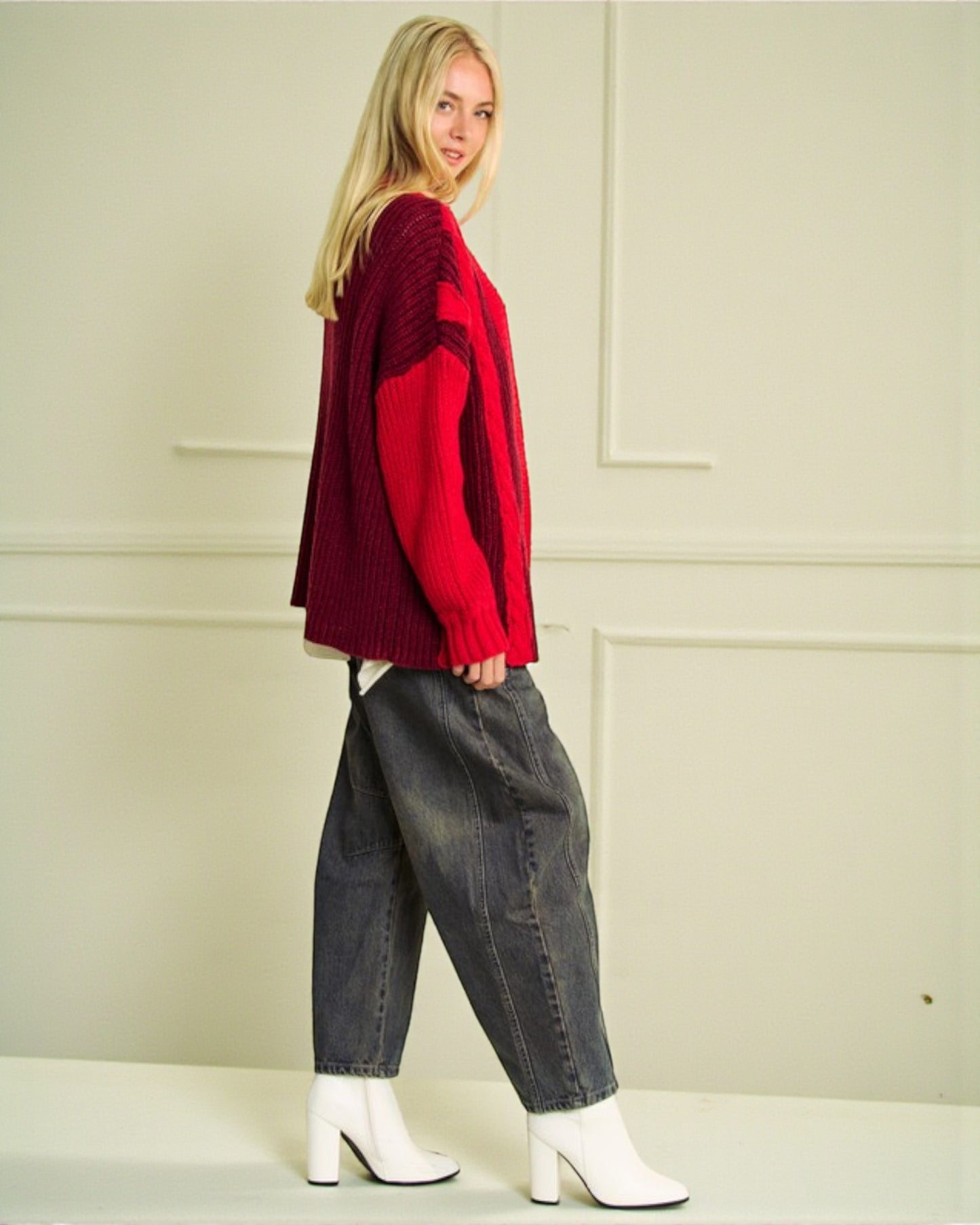 Person wearing a red sweater, dark jeans, and white boots against a light-colored wall.