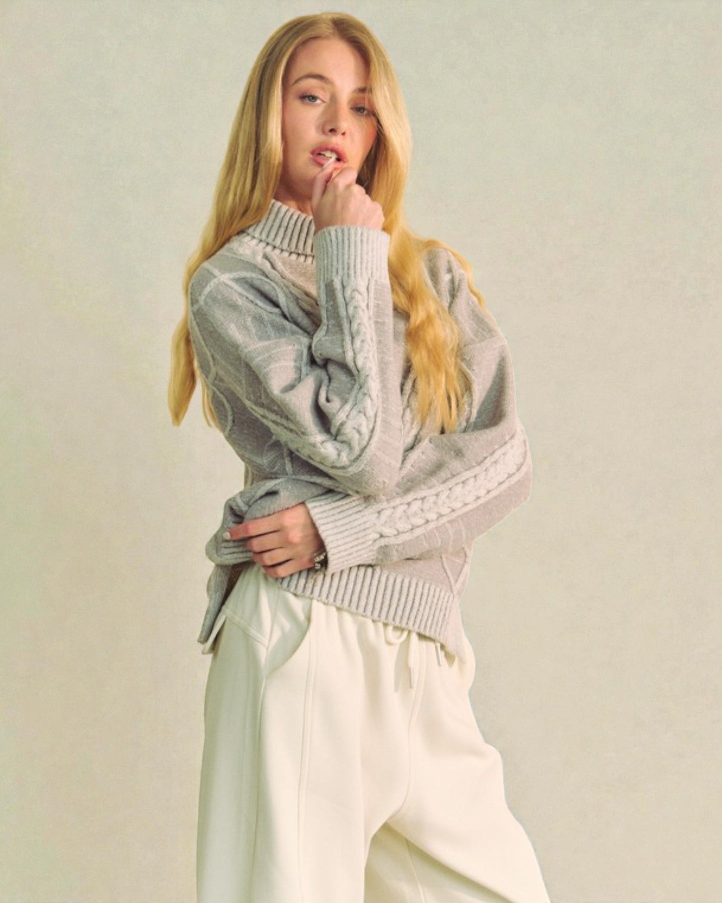 Woman wearing a light gray sweater and white pants against a plain background