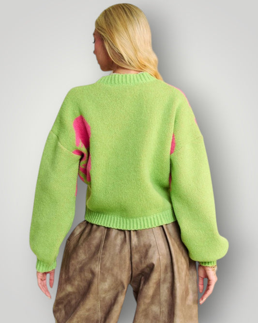 Person wearing a green sweater with pink accents and brown pants on a plain background