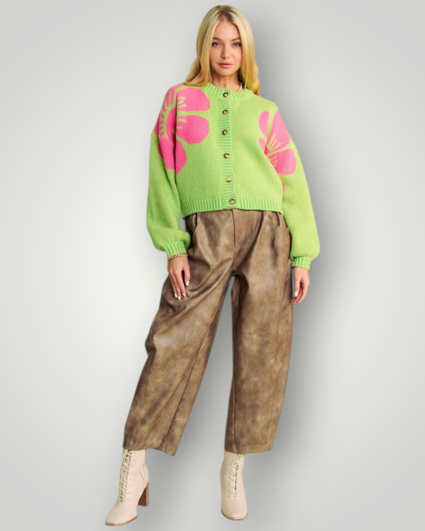 Person wearing a green sweater with pink floral patterns and brown pants on a gray background