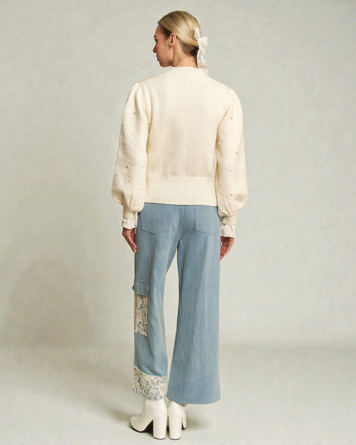 Woman wearing a cream blouse and light blue pants with lace details on a plain background