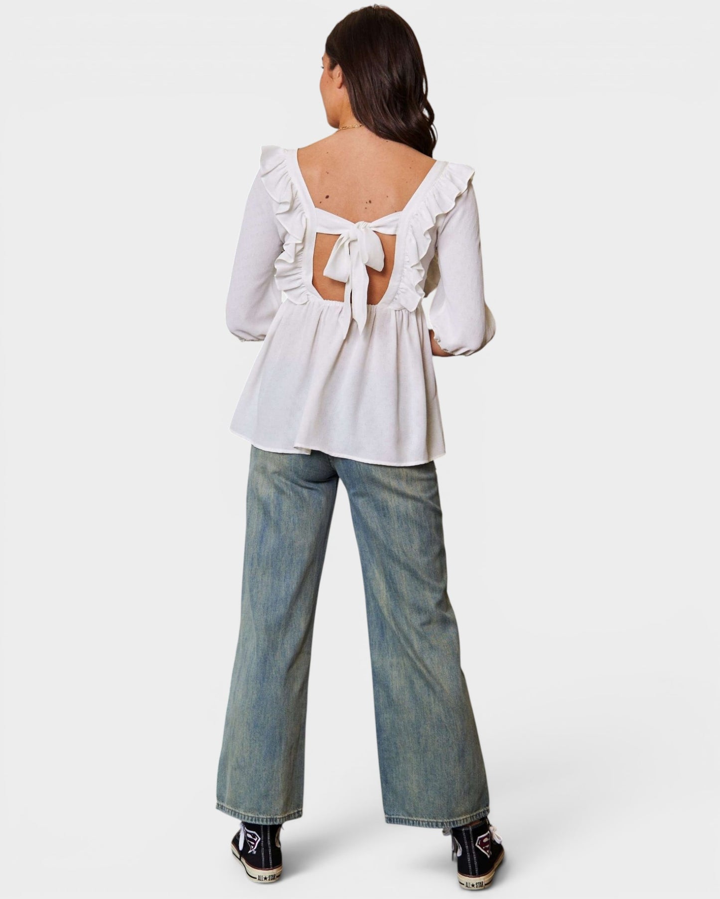 Woman wearing a white blouse with ruffled sleeves and an open back, paired with blue jeans on a white background.