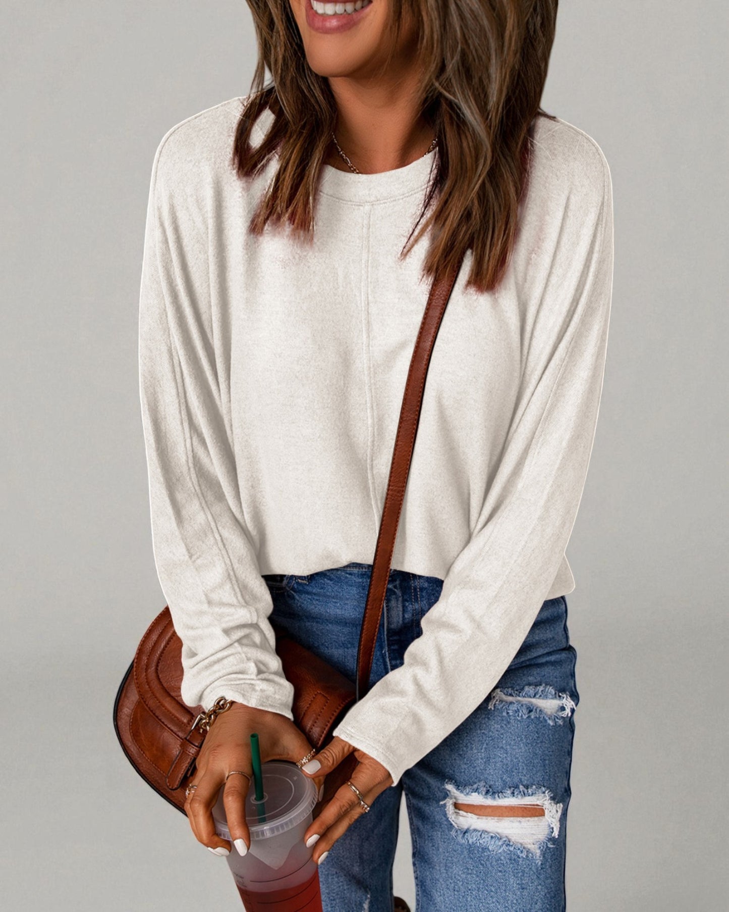 Woman wearing a white long-sleeve top and blue jeans, holding a drink with a straw.