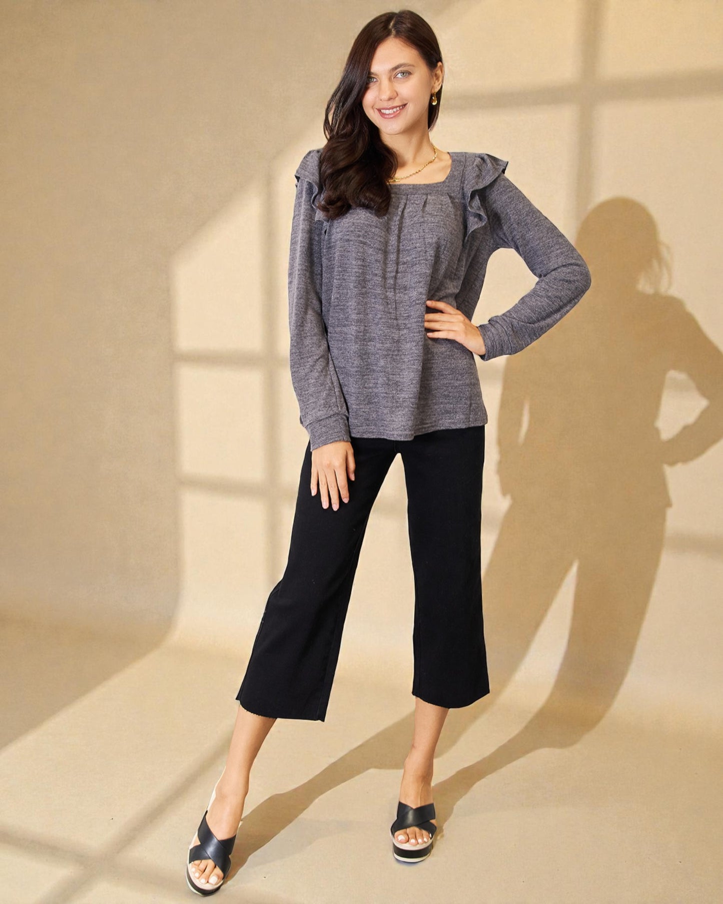 Woman wearing a gray sweater and black pants standing against a beige wall.