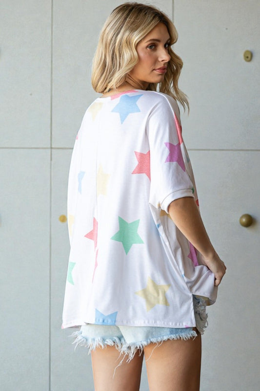 Woman wearing a white oversized t-shirt with colorful star patterns and denim shorts against a light-colored wall.