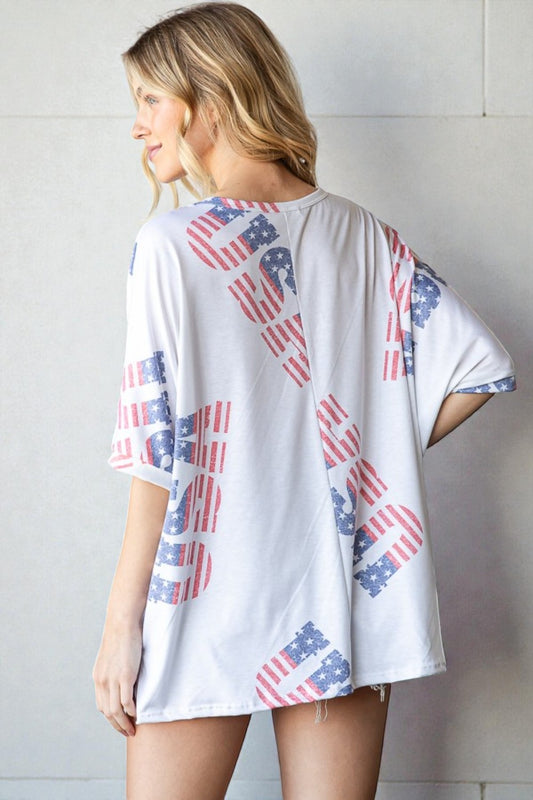 Woman wearing a white t-shirt with American flag patterns on the back and sleeves against a light gray wall.