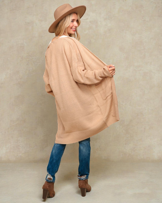 Woman wearing a beige cardigan, blue jeans, and brown boots against a plain background