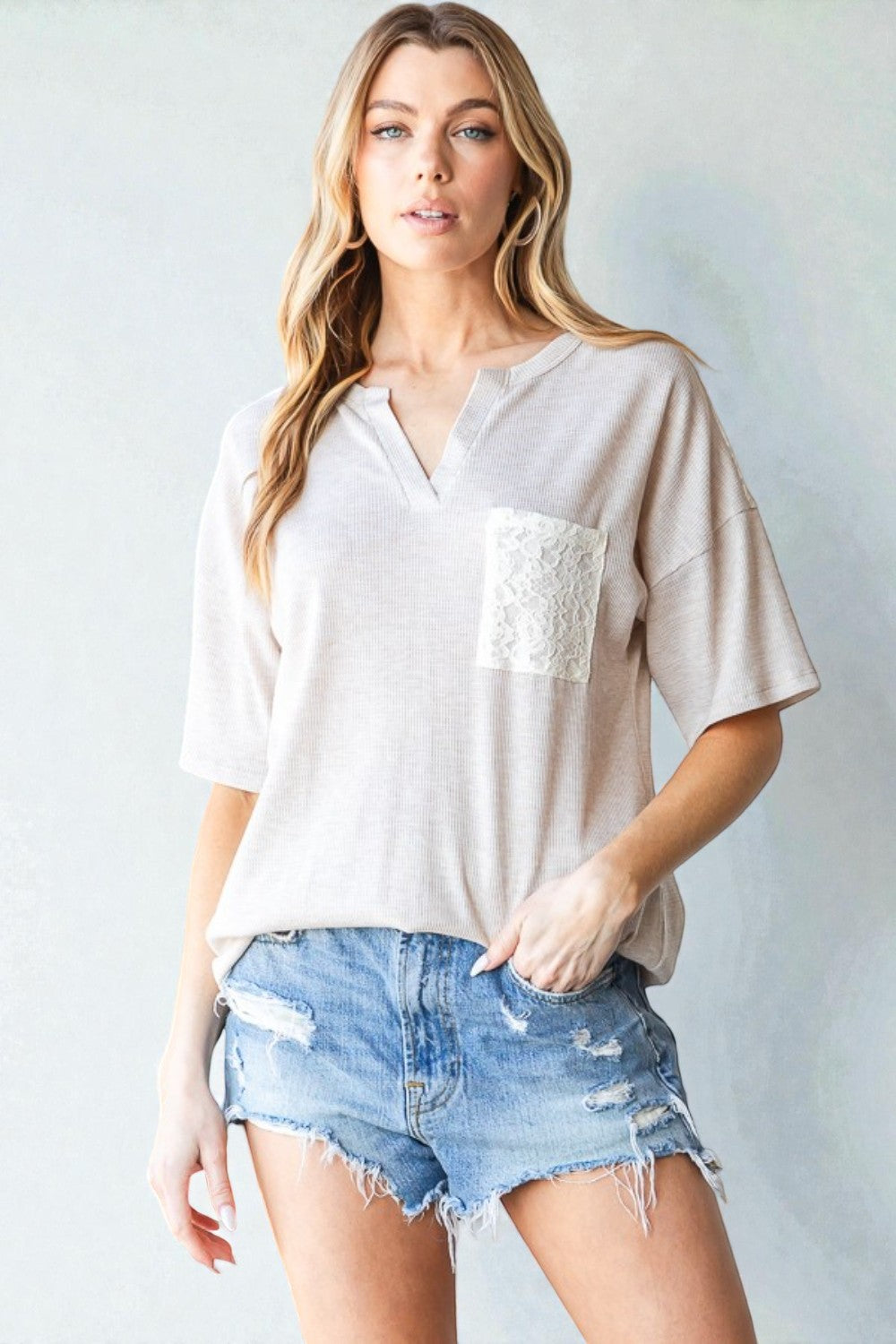 Woman wearing a beige top with lace detail and denim shorts against a plain background