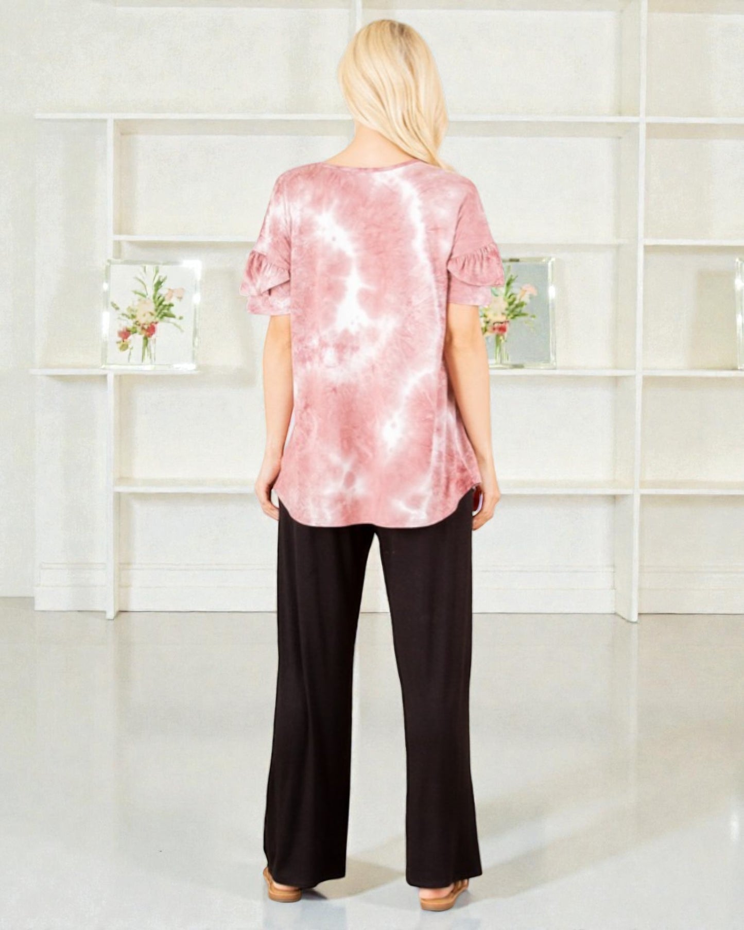 Woman wearing a pink tie-dye top and black pants standing in front of a white shelving unit.