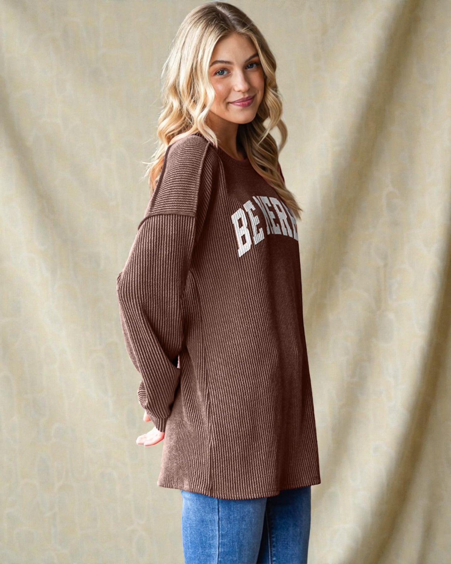 Woman wearing a brown sweater with 'Beverly' text against a beige curtain background