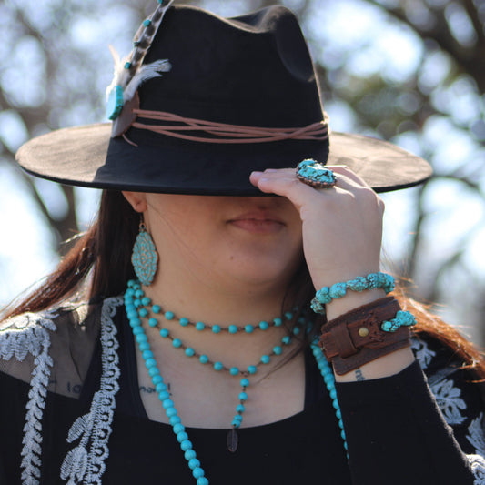 Person wearing a wide-brimmed hat with feathers, turquoise jewelry, and a black top outdoors.