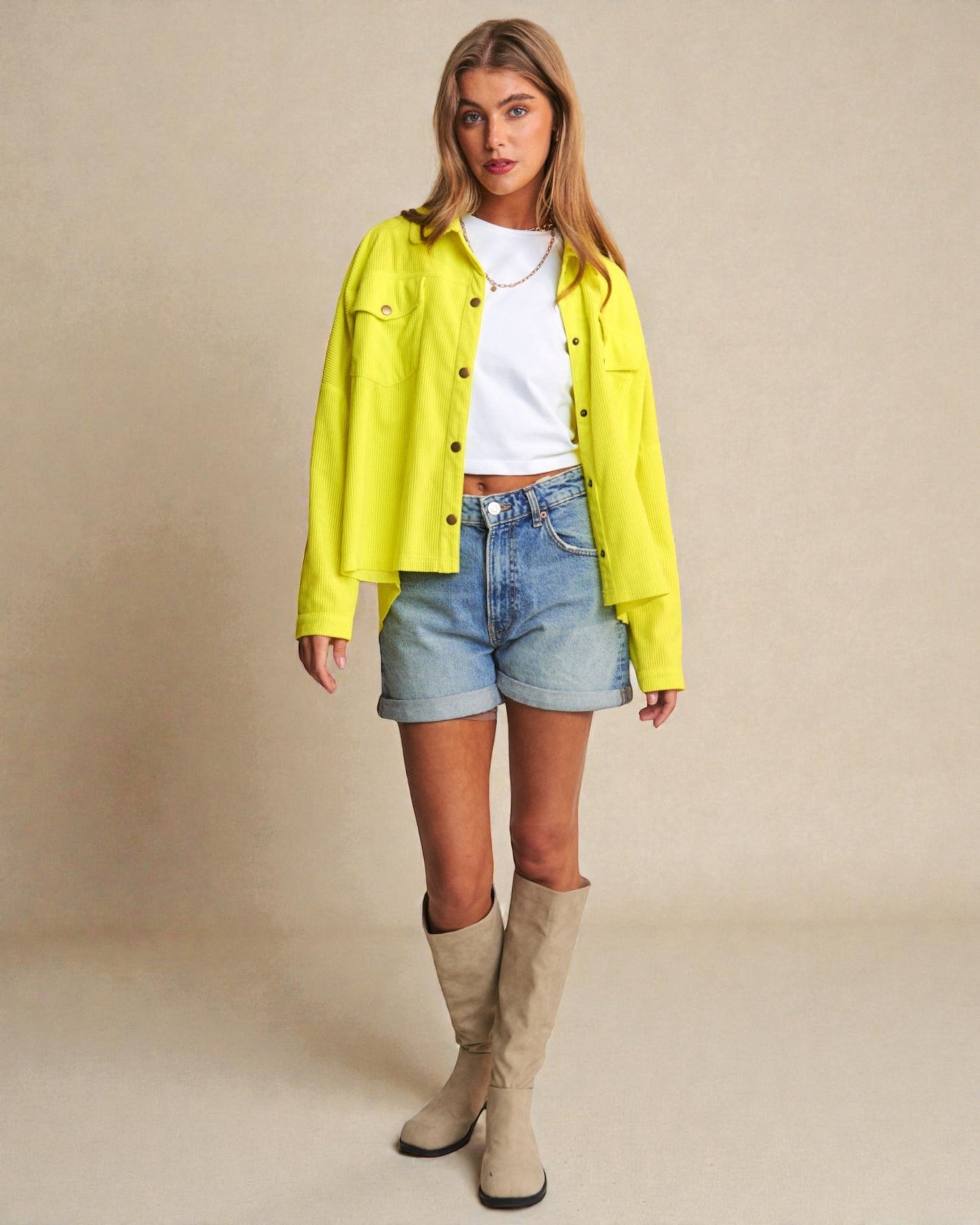 Woman wearing a bright yellow jacket, white shirt, denim shorts, and beige knee-high boots on a beige background