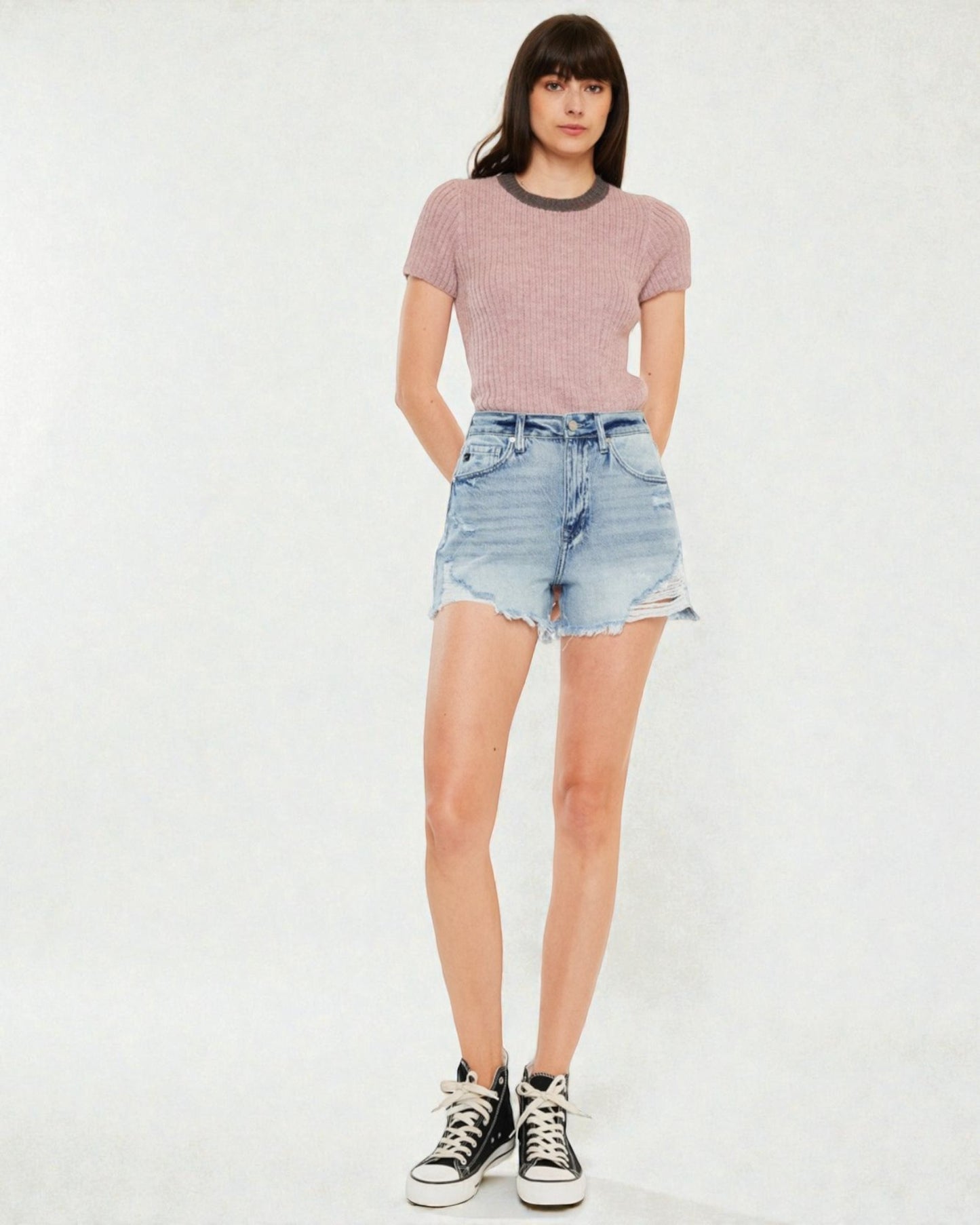 Person wearing a pink ribbed sweater, denim shorts, and black sneakers on a white background