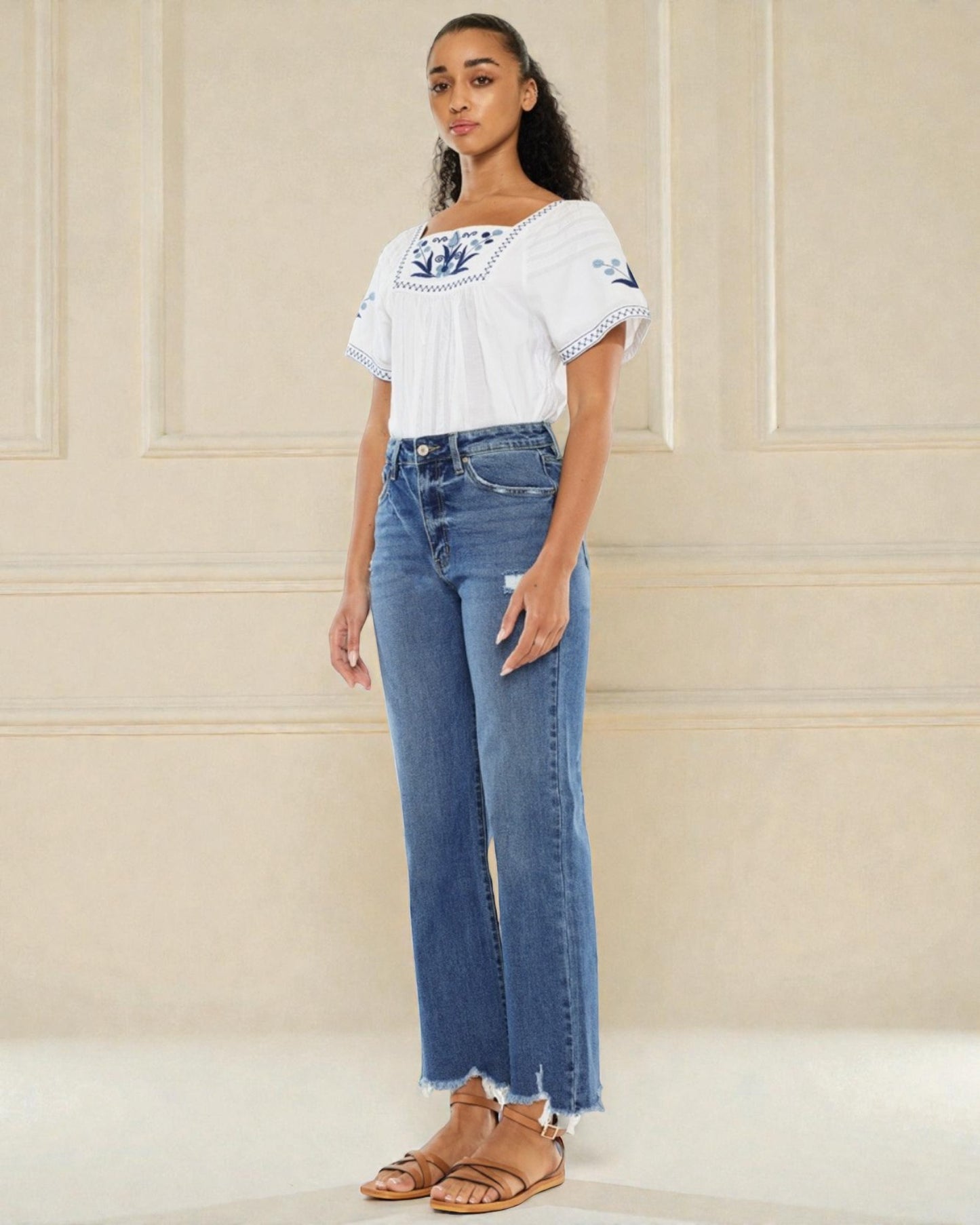 Woman wearing a white blouse with blue patterns and blue jeans against a beige wall.