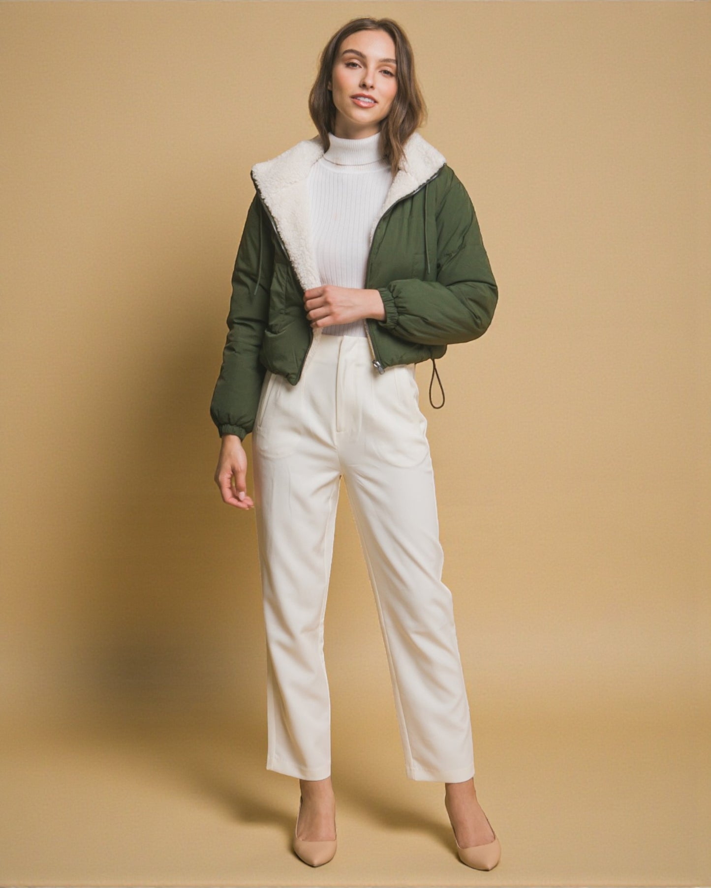 Woman wearing a green puffer jacket with white fur lining, white turtleneck, and white pants on a beige background
