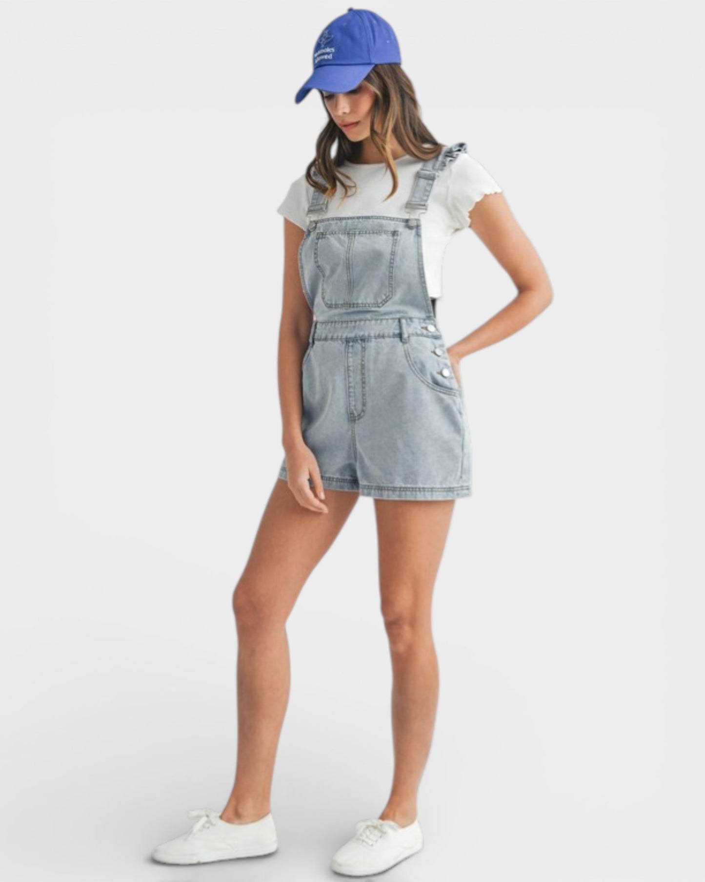 Person wearing a denim overall dress, white sneakers, and a blue cap on a white background
