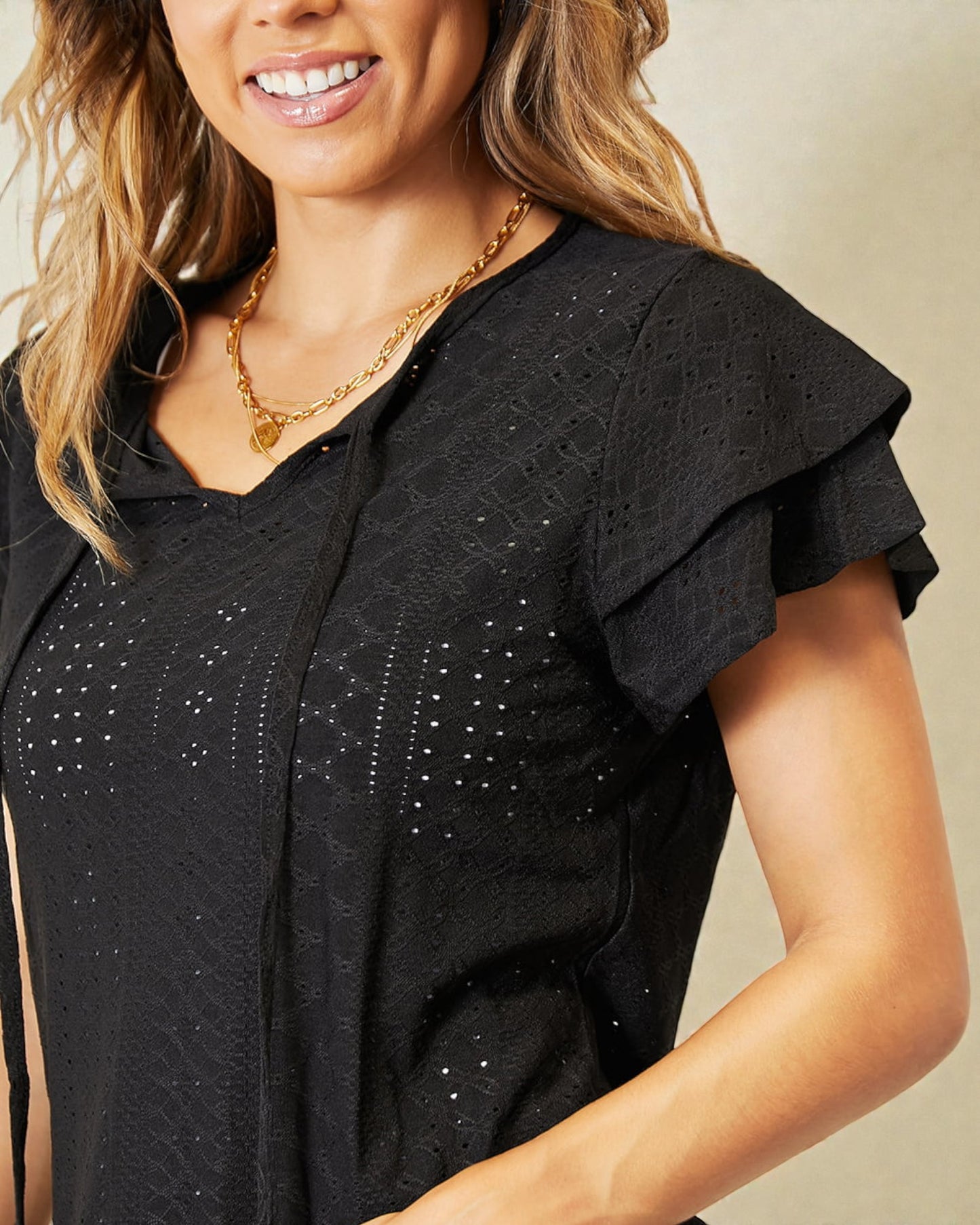 Woman wearing a black top with ruffled shoulders and a gold necklace against a beige background