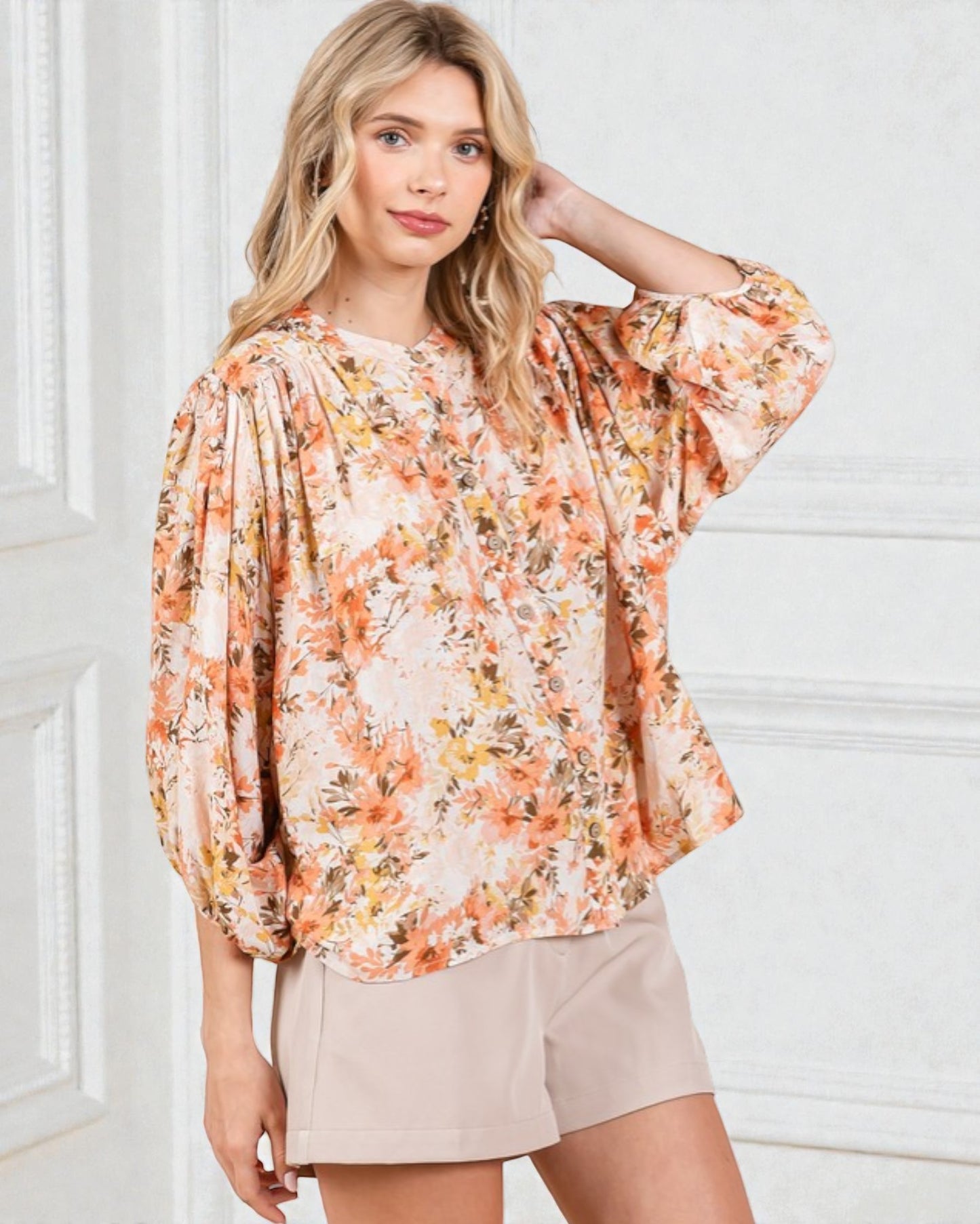 Woman wearing a floral blouse and beige shorts against a white wall