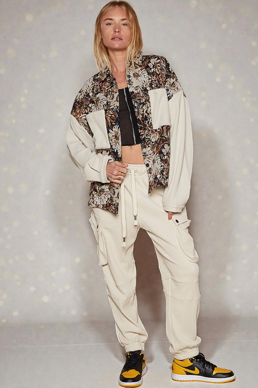 Person wearing a floral jacket, black top, and beige pants with yellow sneakers on a light gray background