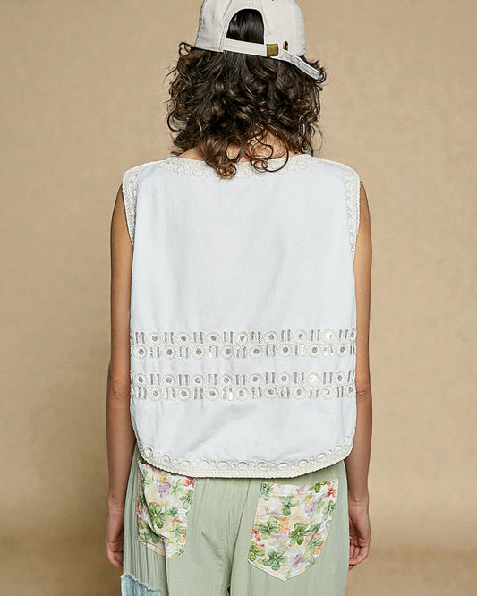 Person wearing a white sleeveless top with floral pocket and light green pants against a beige background