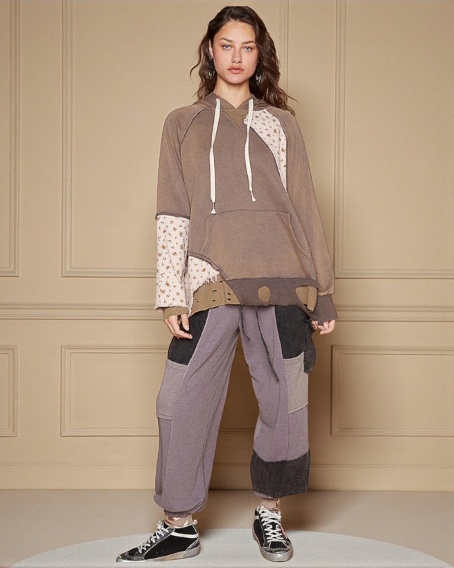 Person wearing a brown hoodie with lace details and gray pants against a beige wall.