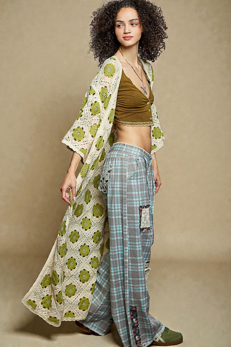 Woman wearing a green patterned top and plaid pants with a long, light-colored shawl over her shoulders against a beige background.