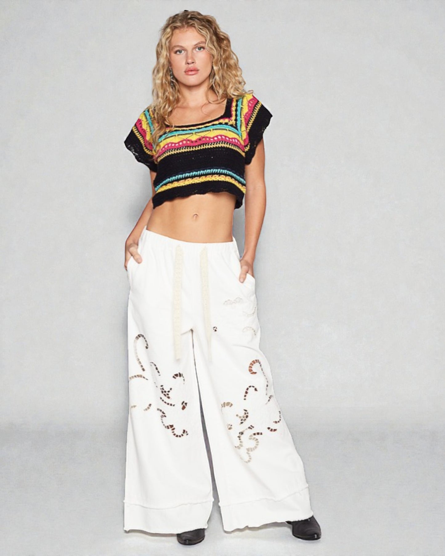 Woman wearing a colorful cropped top and white pants with embroidery on a gray background